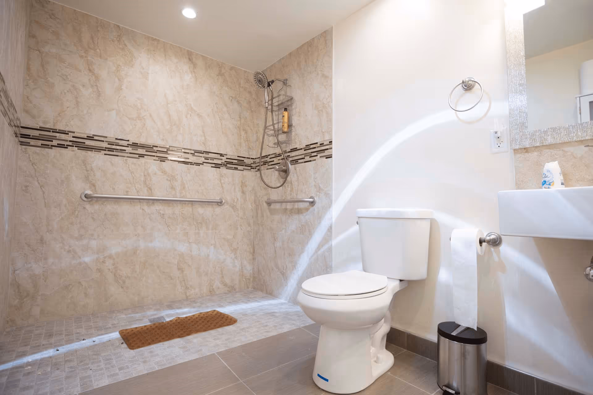 A clean and modern bathroom featuring a walk-in shower with beige marble tiles and a decorative horizontal tile strip. The shower has a handheld showerhead and two grab bars for safety. There is a white toilet next to the shower, a toilet paper holder with a roll of toilet paper, a small trash can, and a sink with a large mirror above it.
