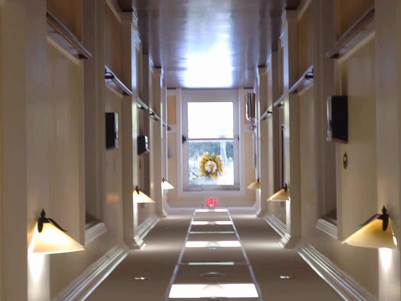 A well-lit hallway in a retirement community with beige walls, wall-mounted lamps on both sides, and a window at the end decorated with a yellow wreath. The hallway has several doorways along each side.