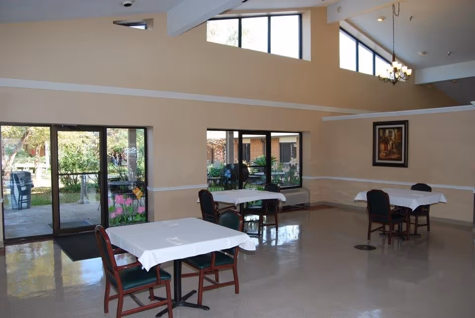 Interior view of a dining area with several tables covered with white tablecloths and green cushioned chairs. Large windows and glass doors provide a view of an outdoor garden area. The walls are beige with white trim, and a framed painting hangs on one wall. A chandelier hangs from the ceiling.