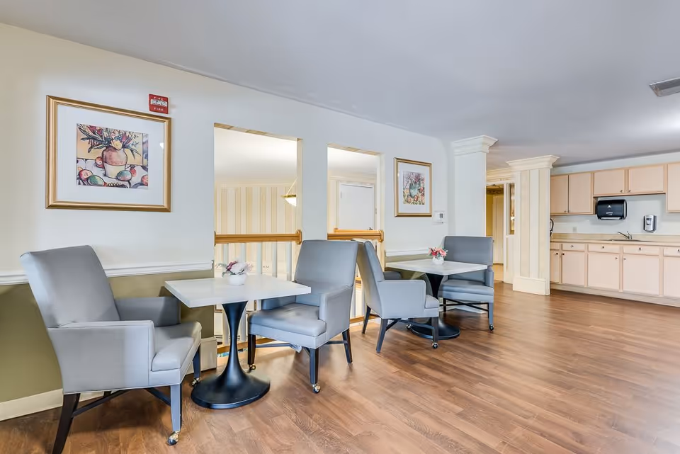 A bright interior room with two small square tables, each accompanied by two gray upholstered chairs on wheels. The room has light-colored walls with framed artwork, wood flooring, and a kitchenette area with cabinets, a sink, and a paper towel dispenser in the background.