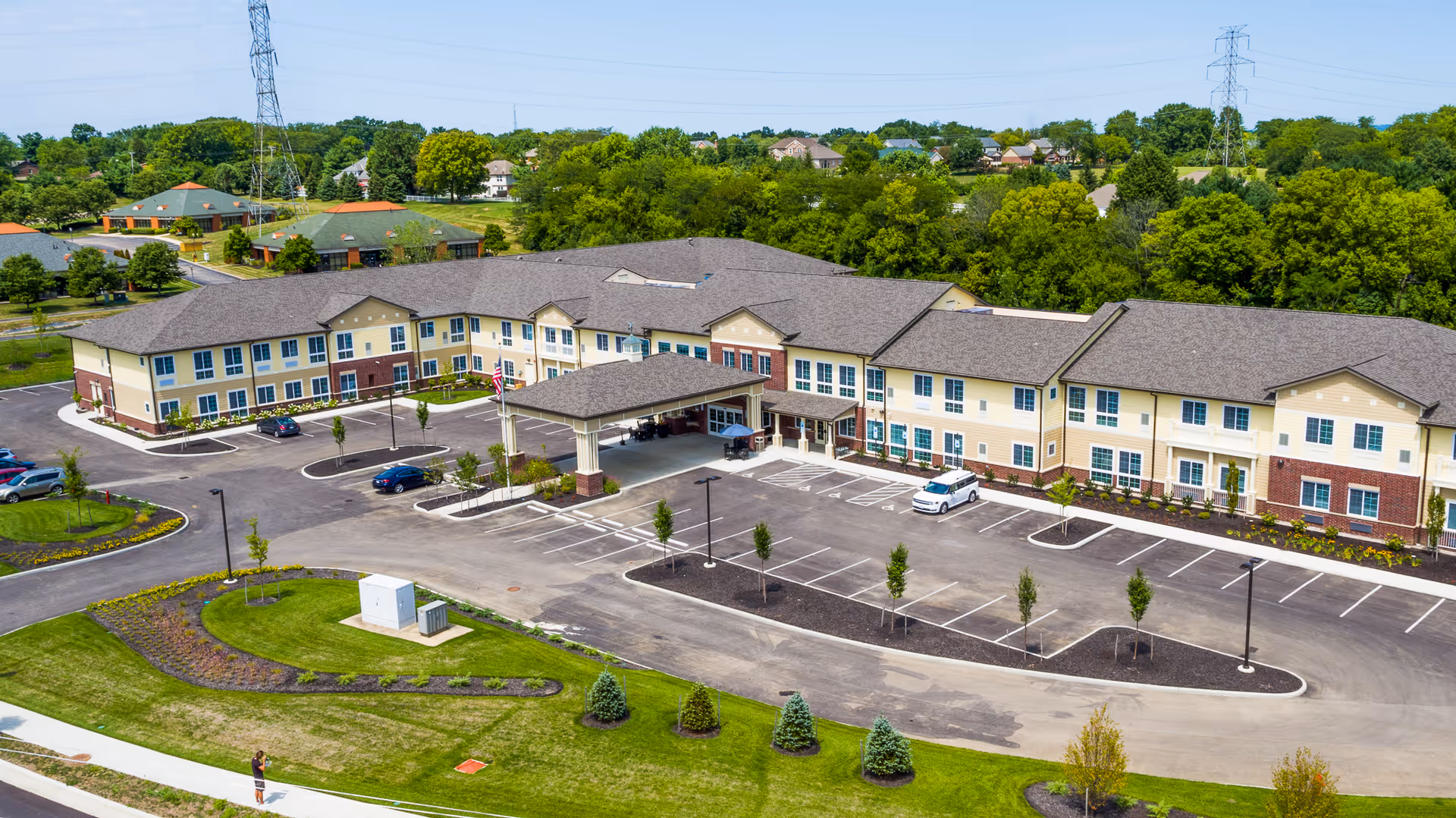 Aerial view of Mason Assisted Living & Memory Care facility showing a large two-story building with a covered entrance, surrounded by parking lots, landscaped greenery, and trees in the background under a clear sky.