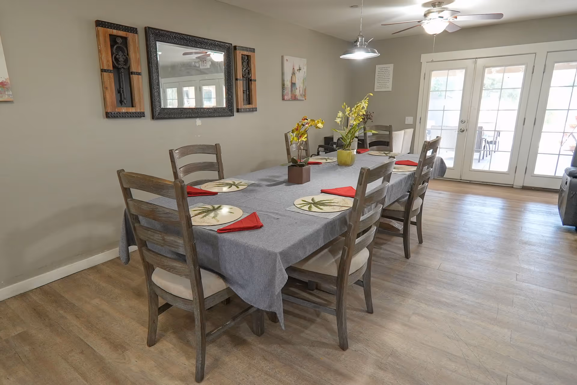 A dining room with a rectangular table covered with a gray tablecloth, set with six chairs. The table has placemats with palm tree designs, red folded napkins, and two flower arrangements. The room has light wood flooring, a ceiling fan with a light, wall decorations including a mirror and artwork, and double glass doors leading outside.