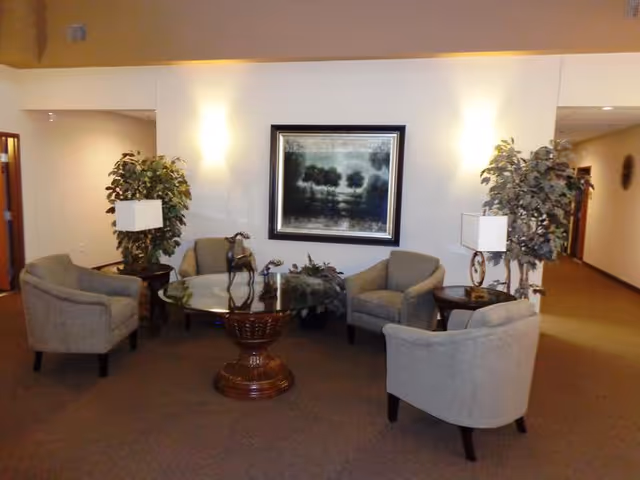 A cozy seating area in a hallway with four beige armchairs arranged around a round glass-top wooden table. The table has decorative items including a small sculpture and greenery. Two side tables with lamps and potted plants flank the seating area. A framed painting of trees hangs on the wall behind the chairs, and the space is softly lit by wall sconces.