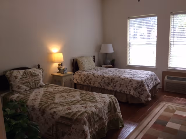 A bedroom with two twin beds covered in floral patterned bedspreads. Between the beds is a small nightstand with a lamp and decorative items. There is a second lamp on a small table near the window. The room has wooden flooring and two windows with blinds letting in natural light. A patterned area rug is partially visible on the floor.