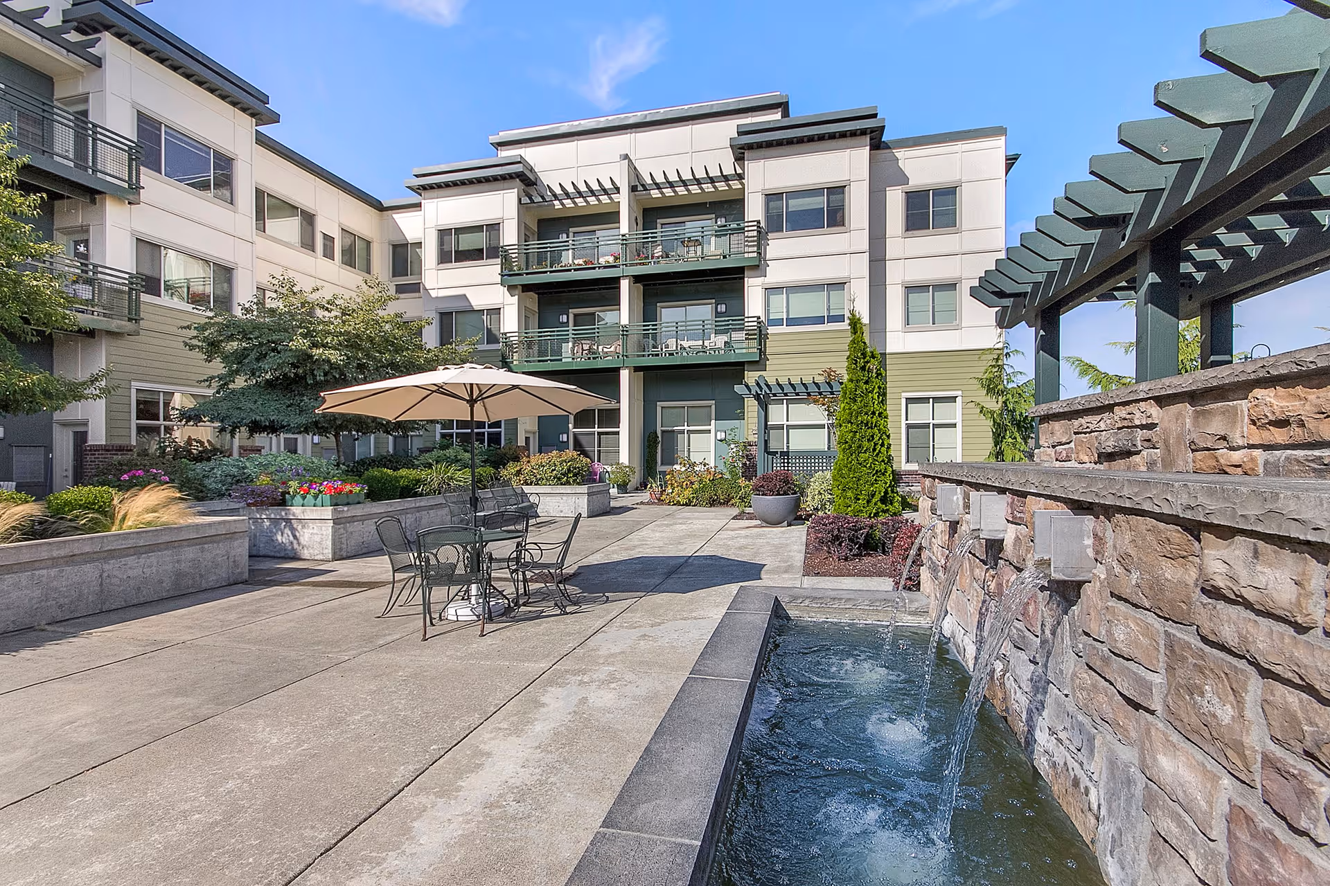 Outdoor courtyard area of a senior living facility with a stone water feature on the right, patio table and chairs with an umbrella in the center, surrounded by landscaped plants and flowers, and a multi-story building with balconies in the background under a clear blue sky.