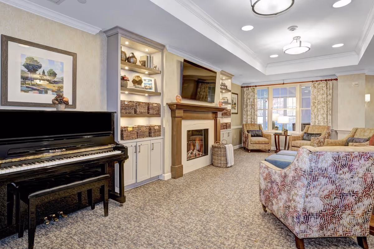 A cozy living room area featuring a black upright piano with a bench, built-in shelves with decorative items and baskets, a fireplace with a mounted flat-screen TV above it, and comfortable upholstered chairs and sofas arranged around a window with patterned curtains.