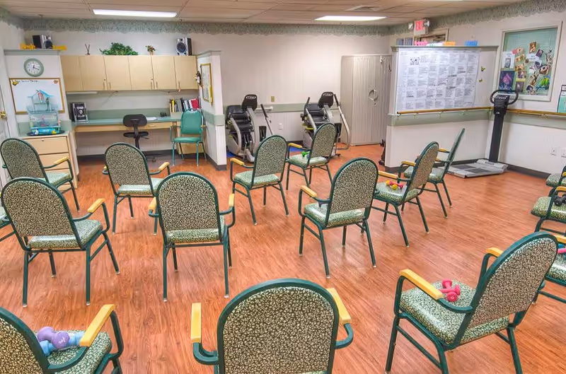 A room with multiple green patterned chairs arranged in a circle on a wooden floor, with small dumbbells placed on some chairs. The room has exercise equipment against the back wall, a bulletin board with papers, a clock, cabinets, and a desk area with chairs.