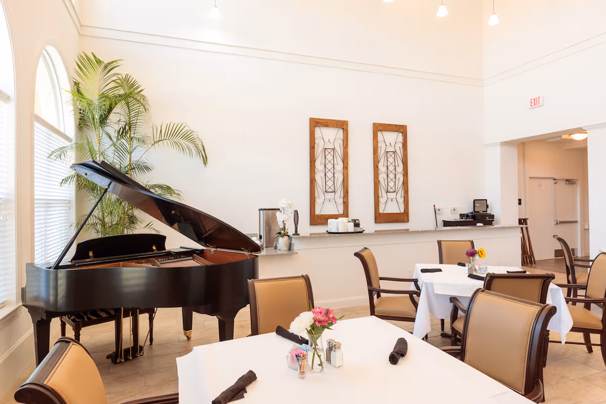 A bright dining room with a grand piano, neatly set tables and chairs, decorative wall art and a service counter.