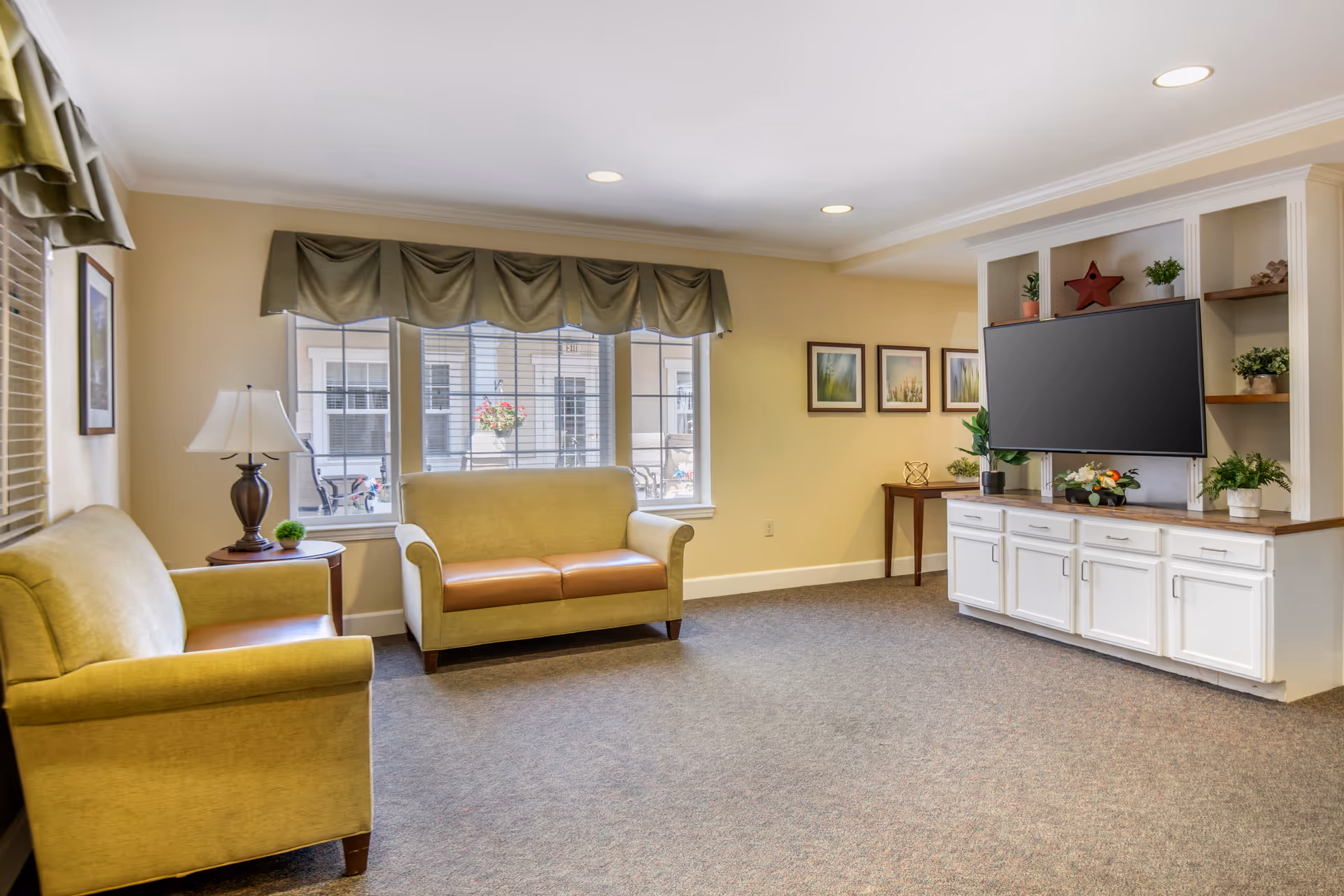 A bright and cozy living room with two beige sofas, a wooden side table with a lamp and a small plant, large windows with green valances, a wall-mounted flat screen TV on a white built-in cabinet with shelves decorated with plants and ornaments, and three framed pictures on the wall.