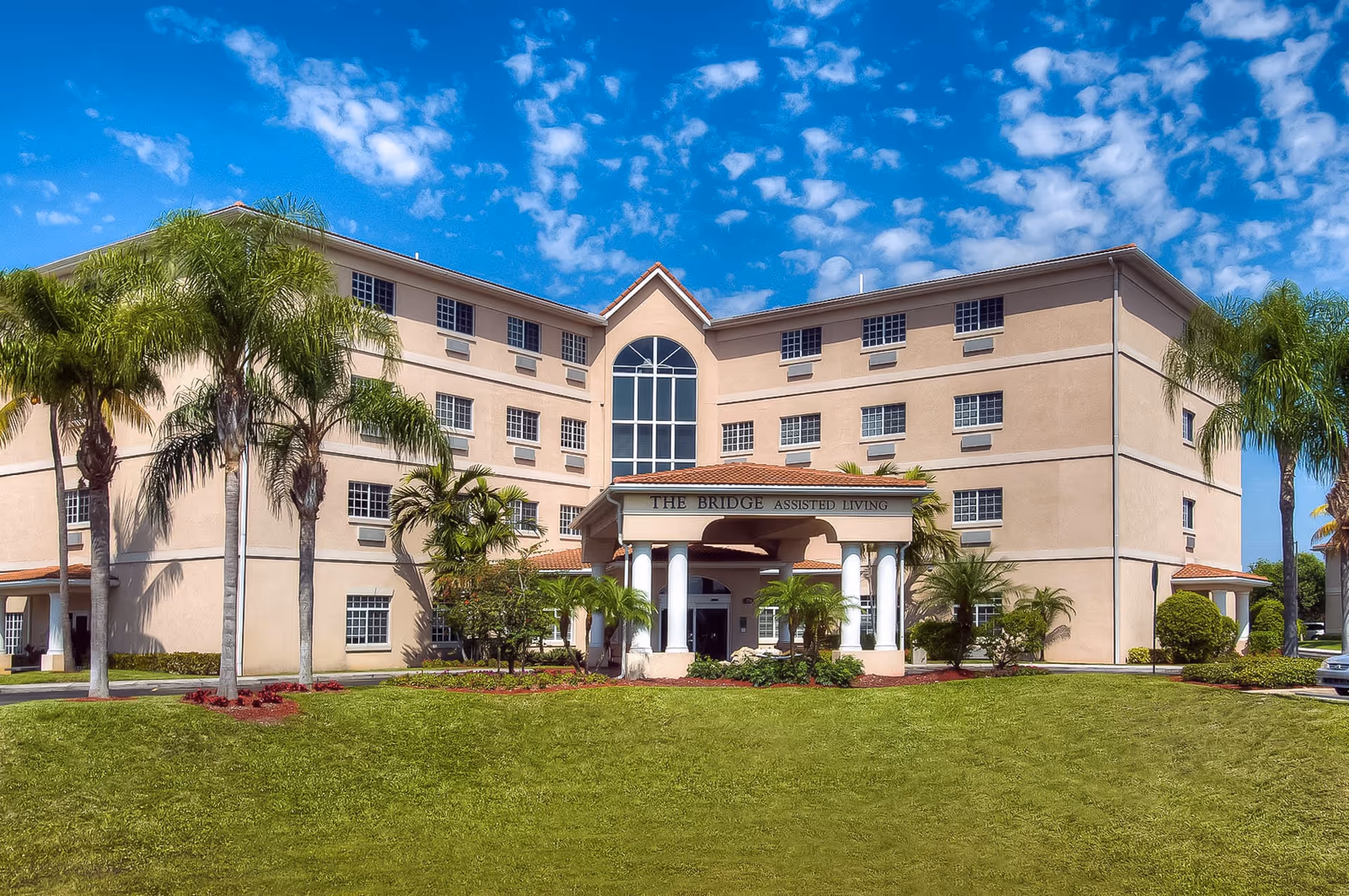 Front exterior view of The Bridge at Inverrary assisted living facility, a beige multi-story building with a red-tiled roof, white columns at the entrance, surrounded by palm trees and green lawn under a partly cloudy blue sky.