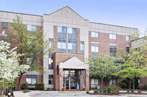 Front view of a multi-story brick senior living facility with glass entrance and trees.