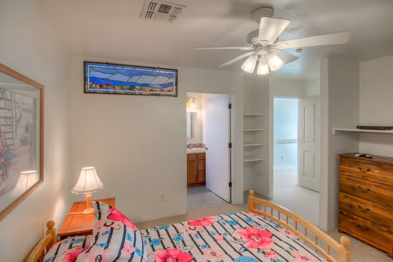 A bedroom with a wooden bed frame and floral bedding in pink, blue, and black. A wooden nightstand with a lamp is beside the bed. On the wall above the bed is a framed picture and a stained glass window. The room has a ceiling fan with lights. There is an open door leading to a bathroom with a vanity and mirror, and another open doorway leading to a hallway with a closet and dresser.