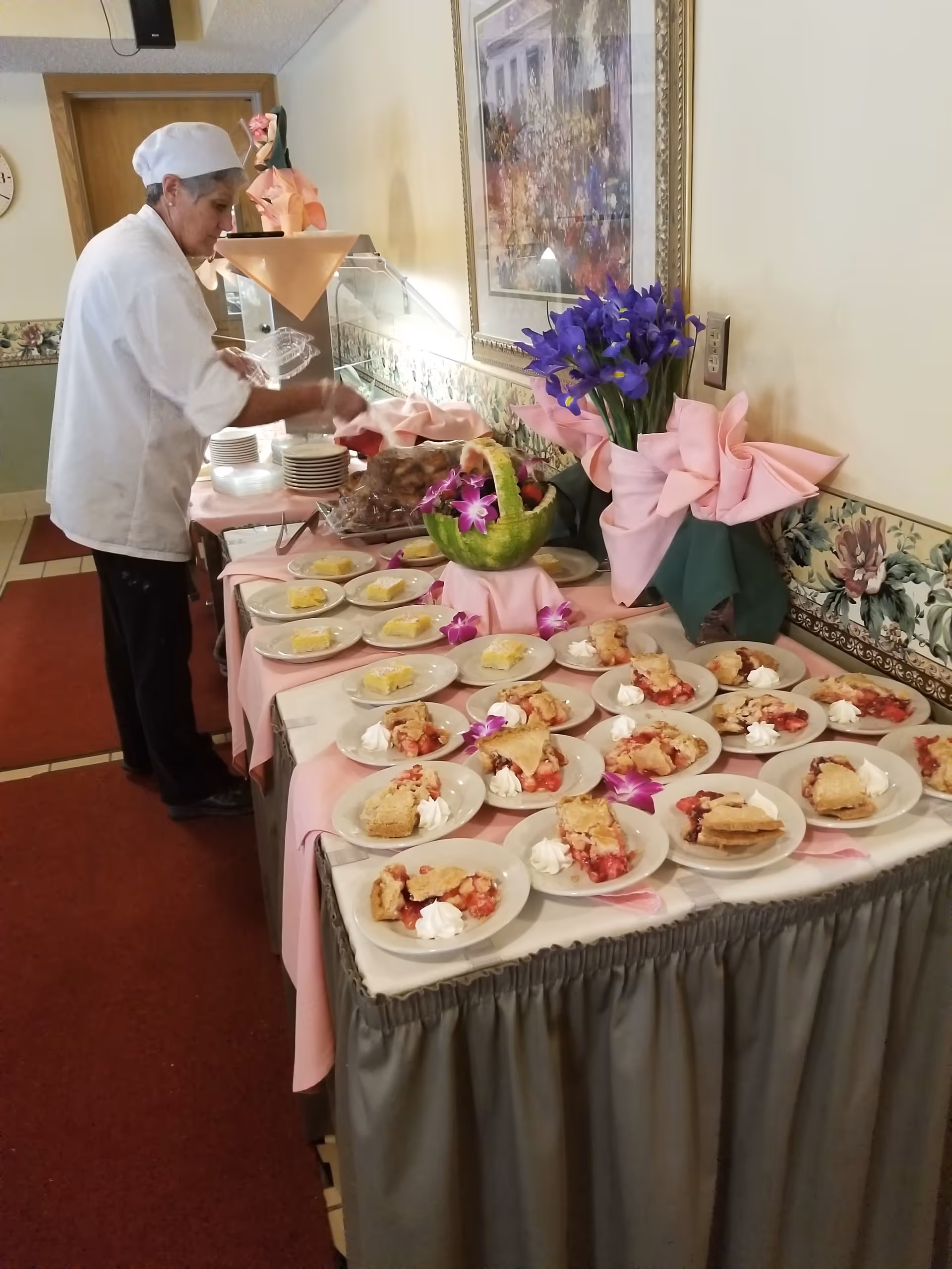 A staff member arranges plates of dessert on a decorated buffet table in a dining area.