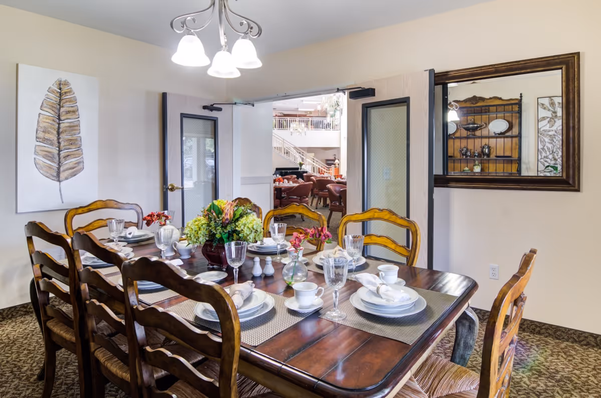 A dining room with a wooden table set for six people, featuring white plates, cups, napkins, and glassware. A floral centerpiece and small vases with flowers decorate the table. The room has beige walls, a carpeted floor, a large mirror on one wall, and a painting of a leaf. Double doors open to a larger common area with additional seating and a staircase.