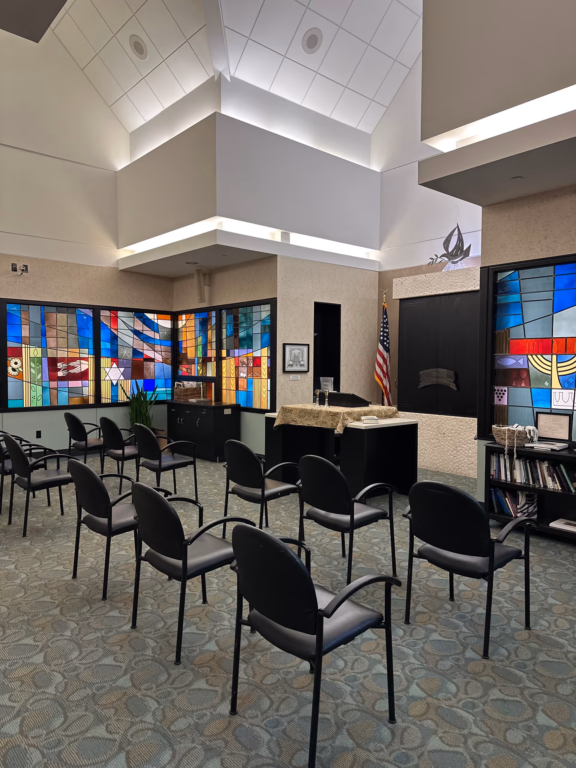 Interior view of a chapel or small assembly room with rows of black chairs arranged facing a podium covered with a cloth. The room features colorful stained glass windows with various designs, an American flag, and a bookshelf with books and framed items. The ceiling is high with modern lighting.