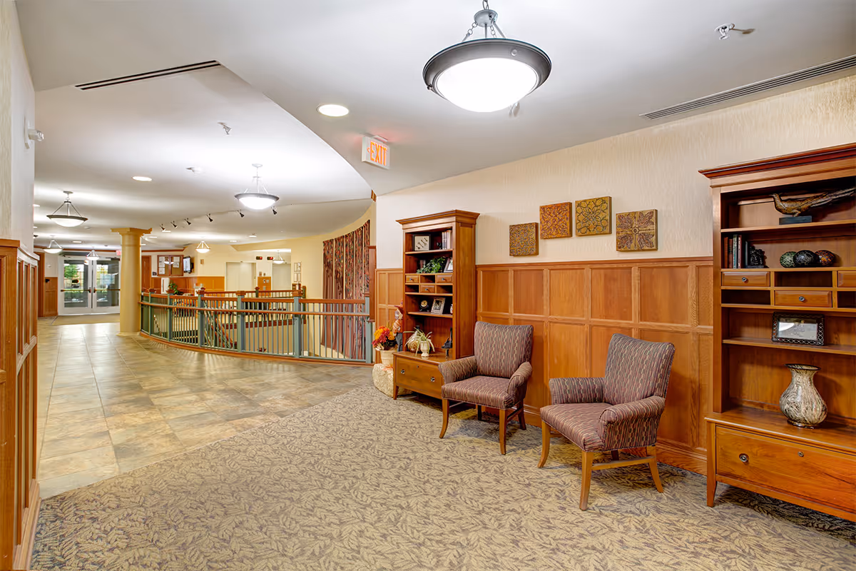 Interior hallway of a senior living facility with wood-paneled walls, two upholstered chairs, wooden bookshelves with decorative items, patterned carpet, tiled floor, ceiling lights, and an exit sign above a door in the background.