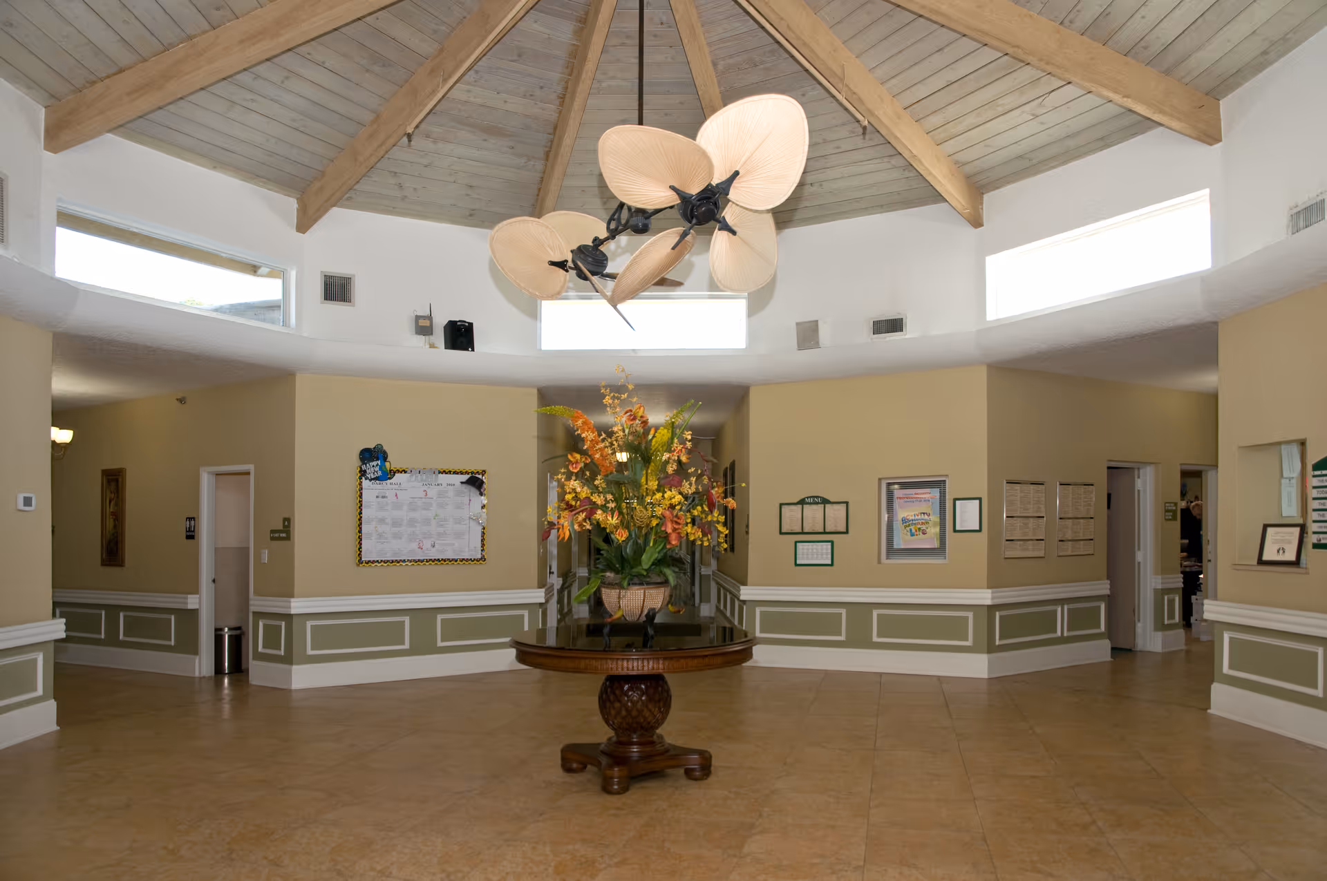 Lobby interior with a round central table topped by a large floral arrangement beneath a wood‑beamed domed ceiling and fan.