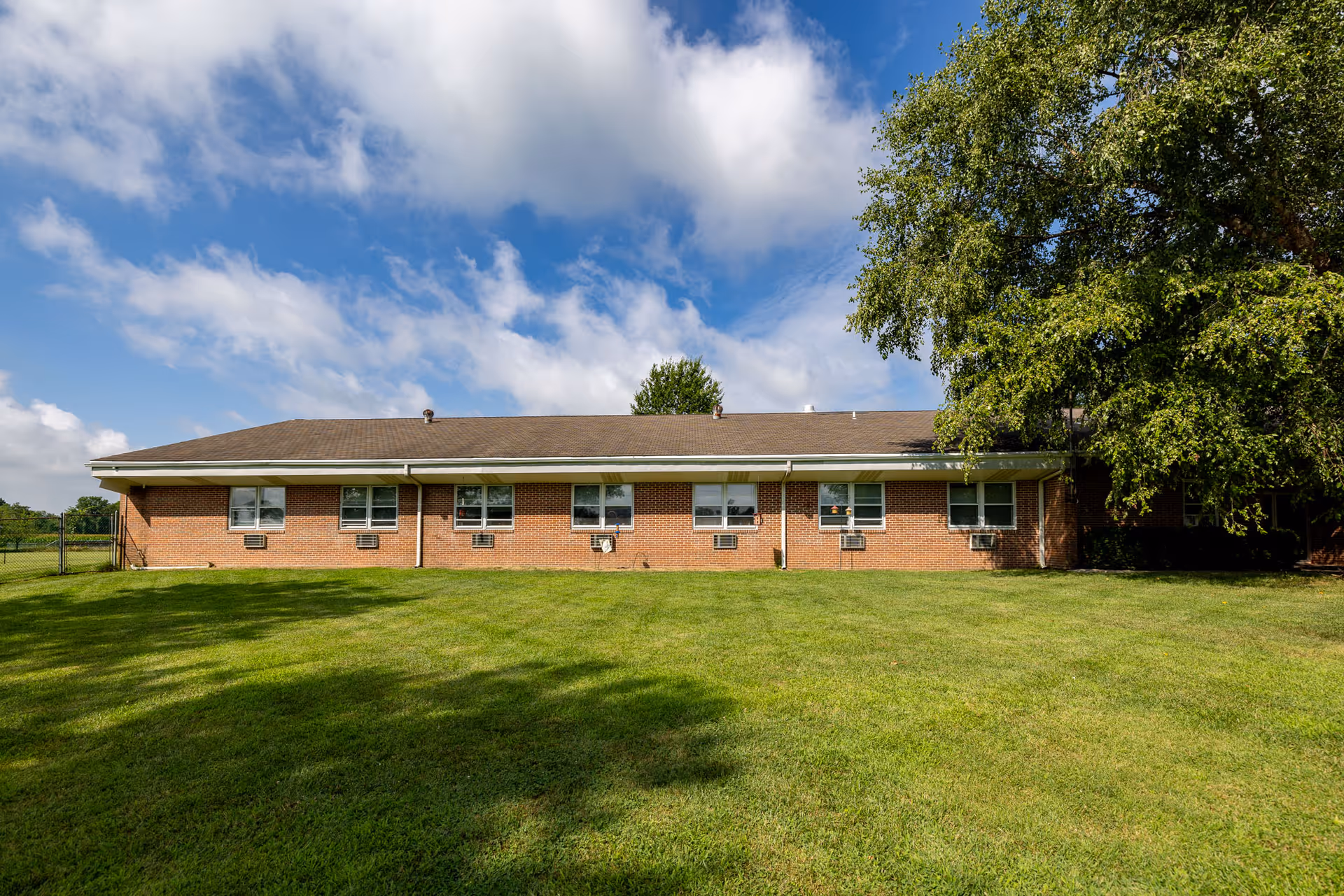Single-story brick building with multiple windows and air conditioning units, surrounded by a large grassy lawn and a tree on the right side under a partly cloudy blue sky.