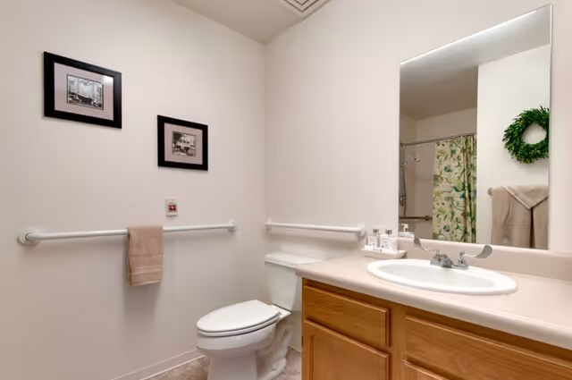 A clean and simple bathroom with a white toilet, a wooden vanity with a white sink, a large mirror above the sink, and a shower with a floral curtain. There are two framed pictures on the wall and a beige towel hanging on a grab bar.