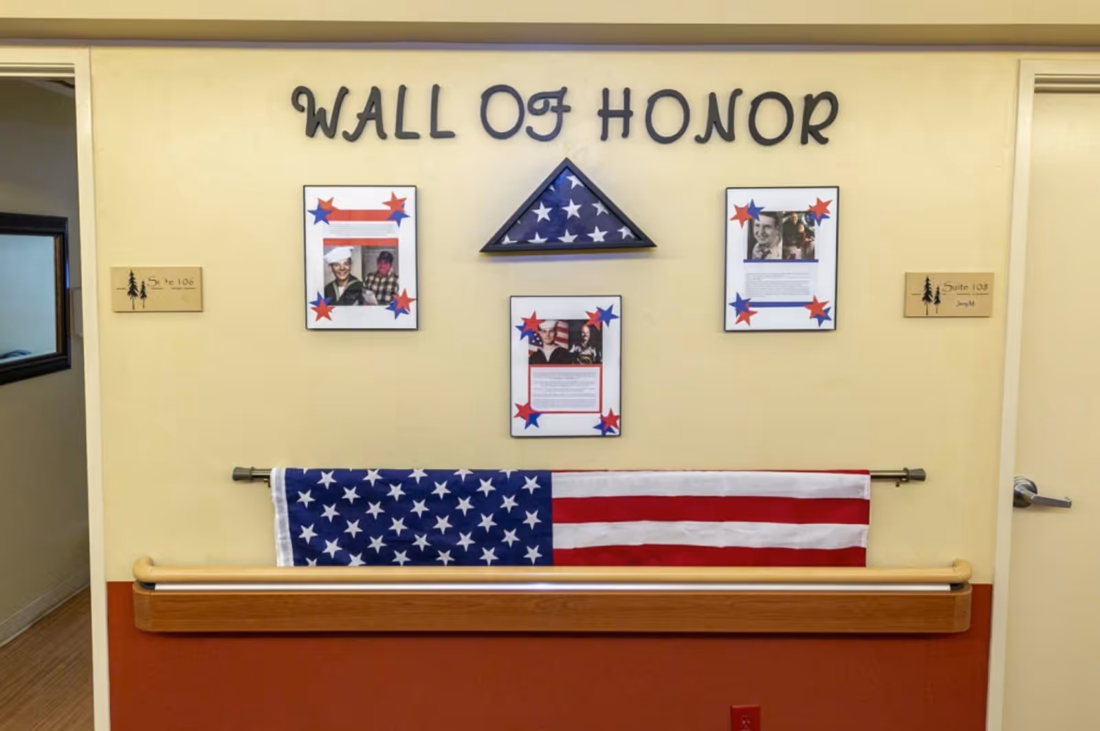 A wall display titled 'Wall of Honor' featuring three framed photos and text, a folded American flag in a triangular display case, and an American flag hanging below. The wall is painted yellow above a red lower section with a wooden handrail. There are two room signs on either side labeled Suite 106 and Suite 108.