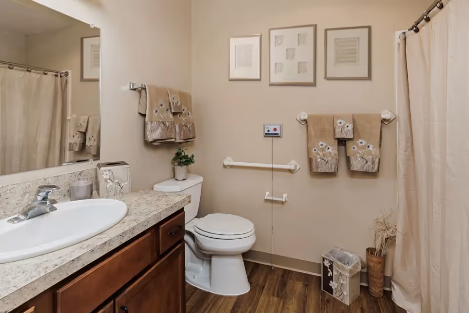 A clean bathroom with a white toilet, a sink with a granite countertop, a large mirror, beige walls, and a shower with a beige curtain. There are decorative beige towels hanging on the wall, a small plant on the toilet tank, framed artwork above the toilet, and a wastebasket on the floor.