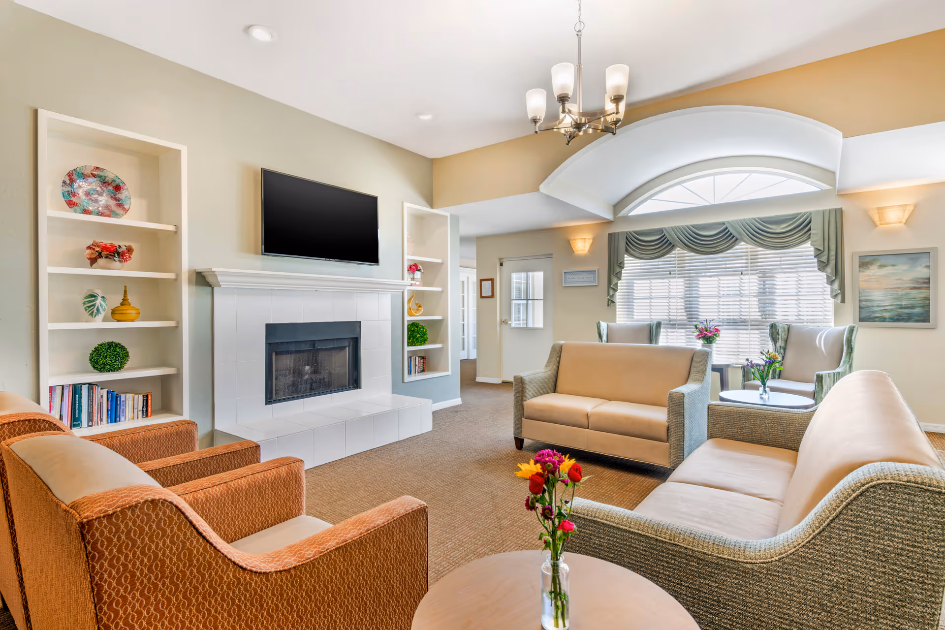 Well-lit common living room with sofas and armchairs arranged around a fireplace and wall-mounted TV, bookshelves, and a large window with drapery.