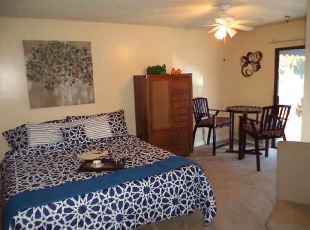A bedroom with a bed covered in a blue and white patterned bedspread and matching pillows. A wooden dresser stands against the wall next to a small round table with two chairs. A ceiling fan with a light is on the ceiling, and a decorative wall hanging is above the bed. A glass door lets in natural light from outside.
