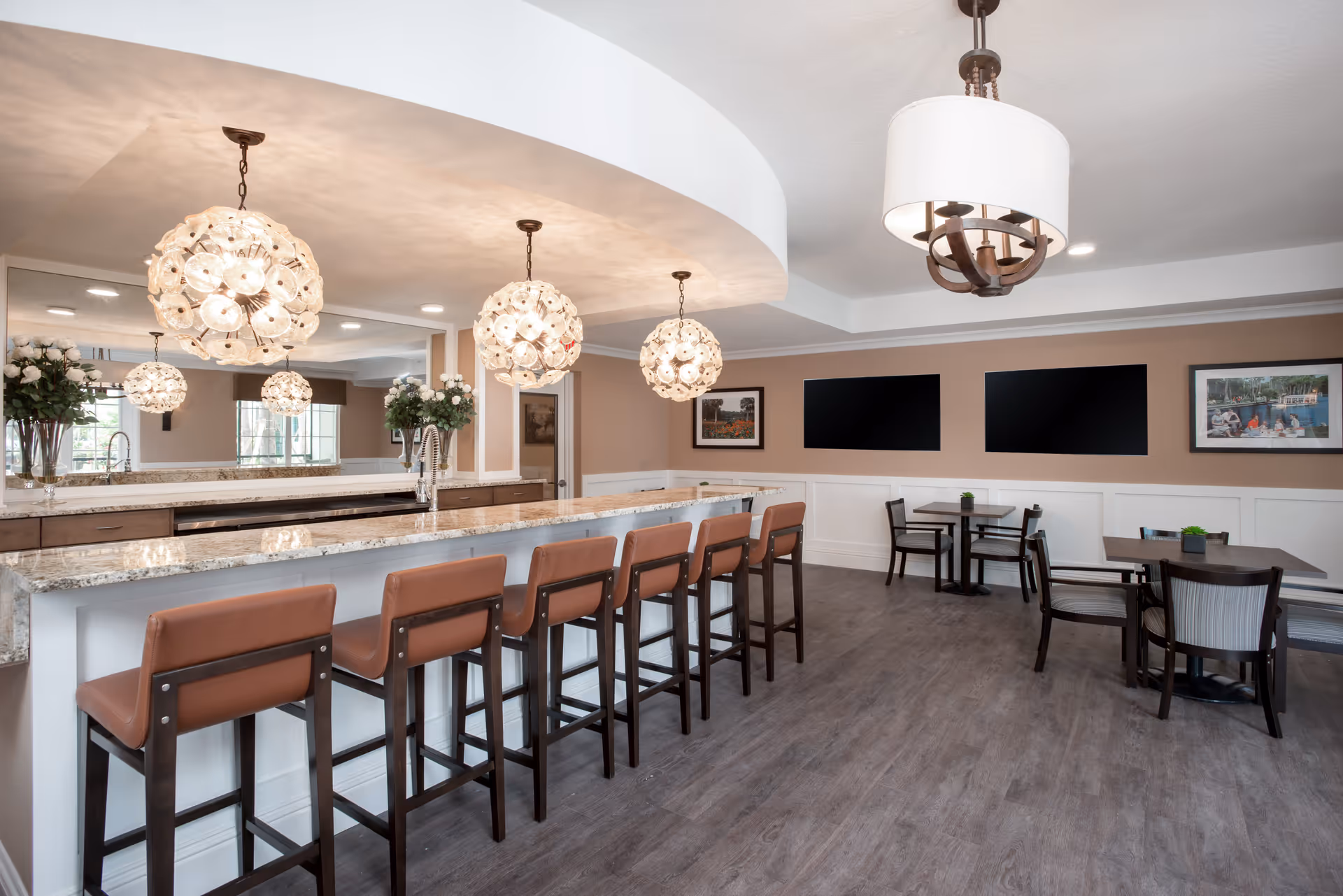 A modern dining area featuring a long granite countertop with seven brown cushioned bar stools. Above the counter hang three decorative pendant lights with floral designs. The room also includes several small dining tables with chairs, framed pictures on the walls, and a large mirror behind the counter reflecting the space.