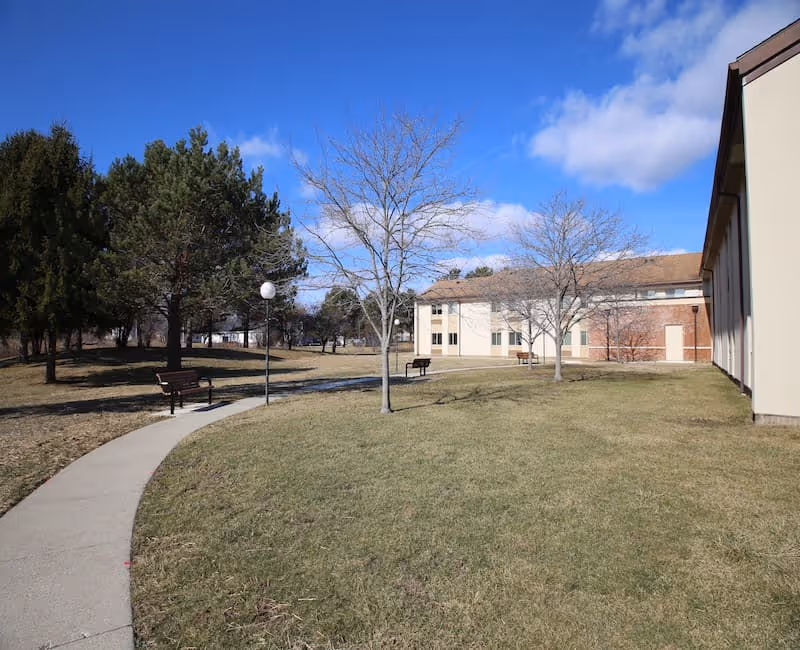 Outdoor view of a senior living facility with a curved concrete pathway, benches, leafless trees, and a building with beige and brick walls under a blue sky with some clouds.