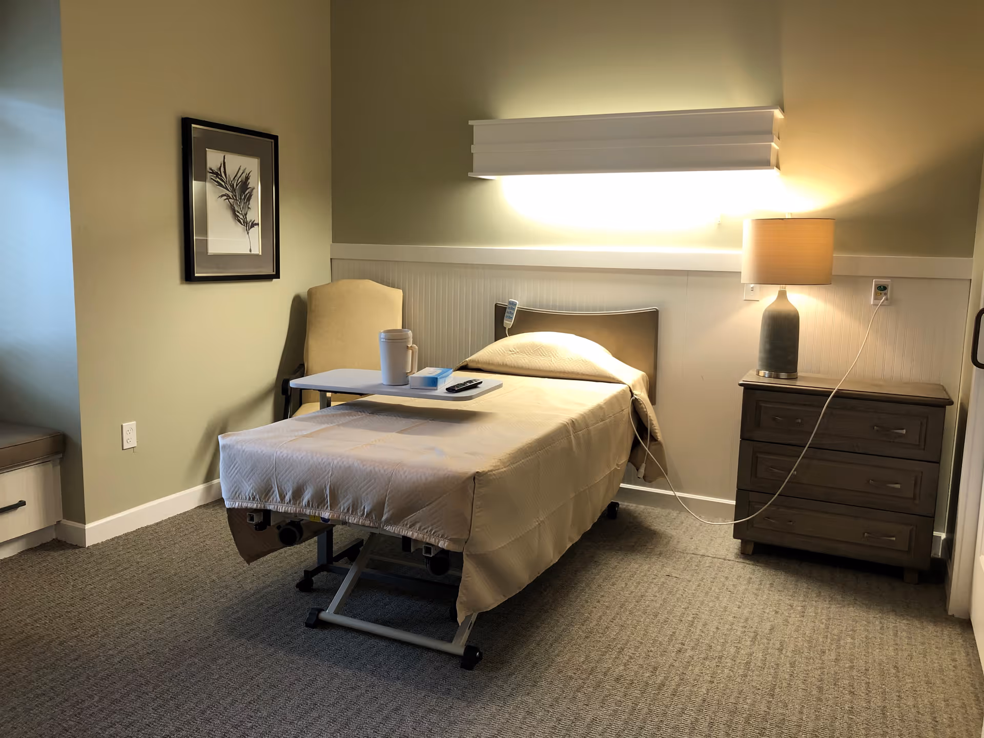 A simple, clean bedroom in a senior living facility with a single bed covered in beige bedding. There is a beige armchair next to the bed, a small overbed table with a cup and remote control on it, a nightstand with a lamp, and a framed botanical print on the wall. The room has soft lighting and neutral-colored walls and carpet.