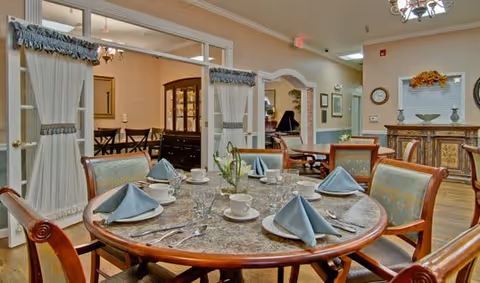 A round dining table set with blue folded napkins, white cups, glasses, and silverware in a well-lit dining area with wooden chairs. In the background, there is a china cabinet and a hallway leading to another room with additional seating and decor.