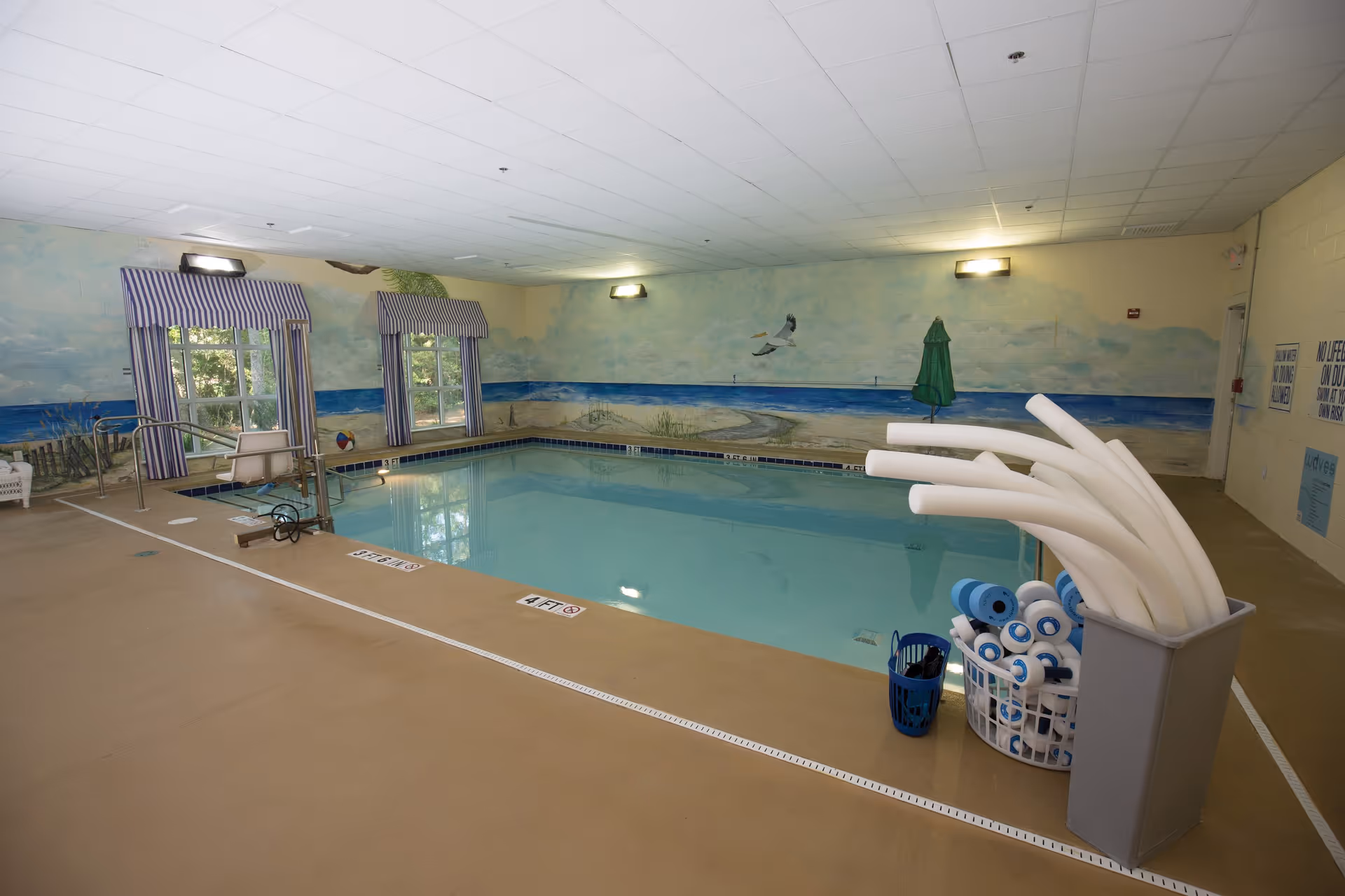 Indoor swimming pool with a seaside mural, pool lift, striped window valances, and baskets of pool noodles and flotation noodles.