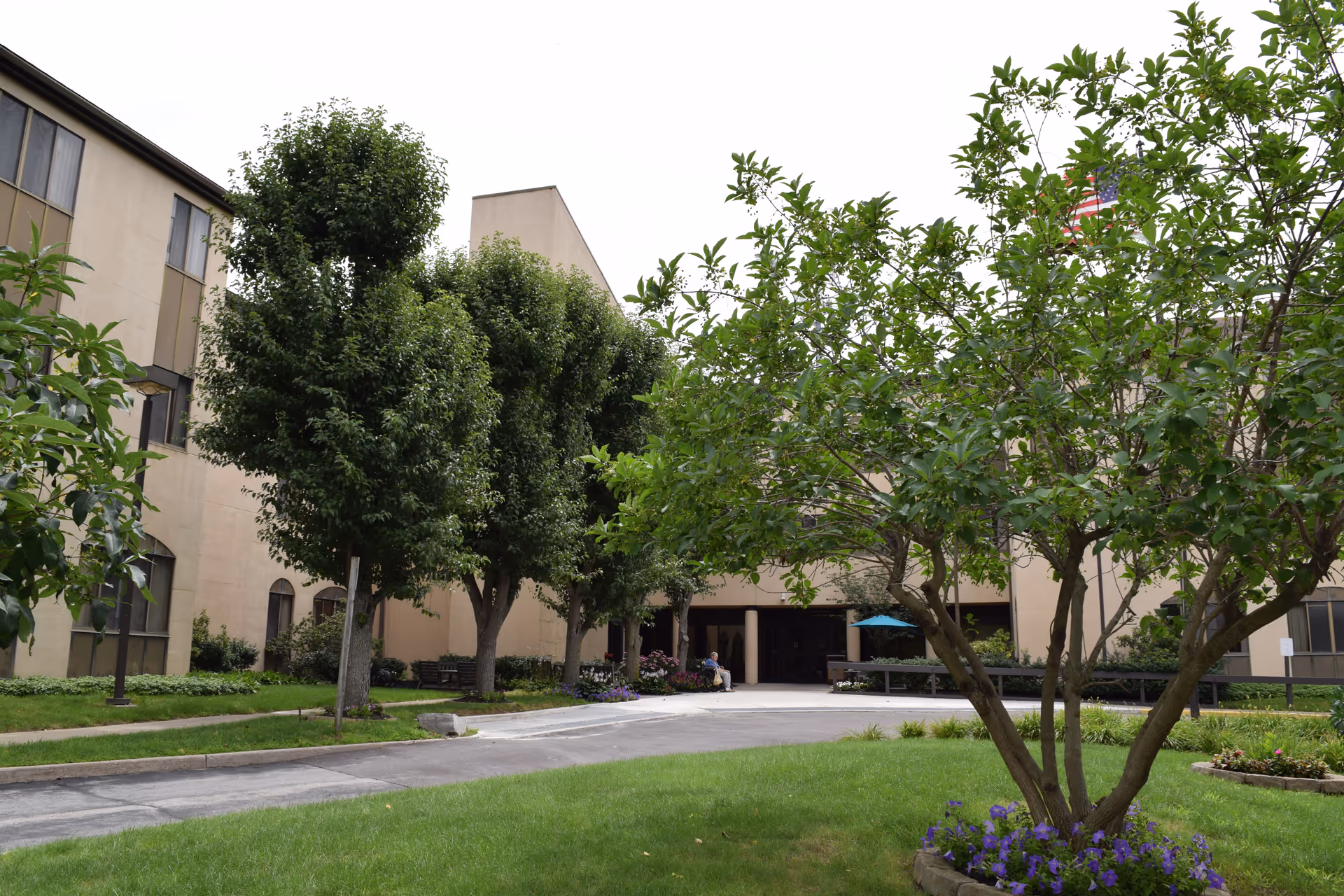 Front courtyard and entrance of a multi-story senior living building with trees, lawn, and a driveway.