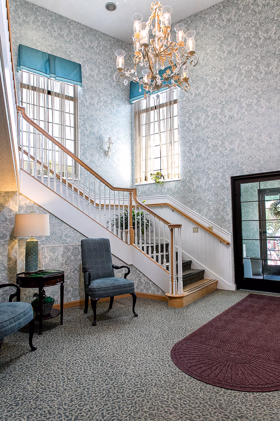 Interior view of a senior living facility lobby area with a staircase featuring wooden handrails and white balusters. The walls are covered in patterned wallpaper, and two tall windows with teal valances allow natural light to enter. A chandelier hangs from the ceiling. Two upholstered chairs and a small round table with a lamp are placed near the staircase. A maroon rug lies on the carpeted floor near a glass door entrance.