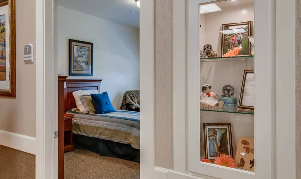 View through a doorway into a bedroom with a neatly made bed featuring multiple pillows, a wooden headboard, and a framed picture on the wall. To the right, a built-in glass display cabinet contains framed photos, decorative flowers, and small ornaments.