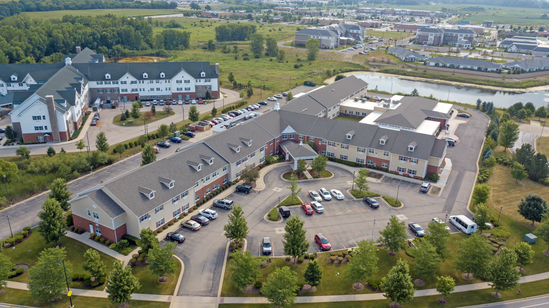 Aerial view of Heritage Woods of Huntley senior living facility showing multiple connected buildings with gray roofs, surrounded by parking lots, green lawns, trees, and a small pond in the background.