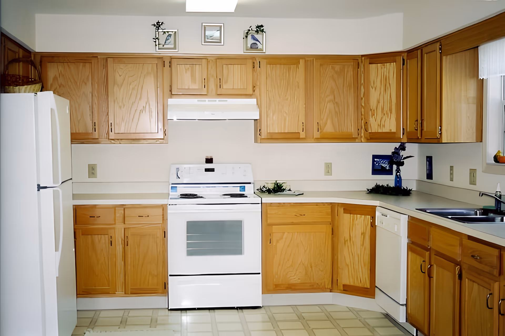 A kitchen with wooden cabinets, a white refrigerator on the left, a white stove with an oven in the center, and a white dishwasher on the right. The countertops are light-colored, and there is a window with a white curtain above the sink on the right side. Small decorative items and framed pictures are placed above the cabinets.
