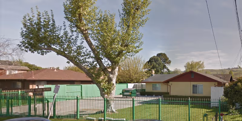 Single-story ranch-style buildings behind a green metal fence and gate, with a large tree and lawn in the front.