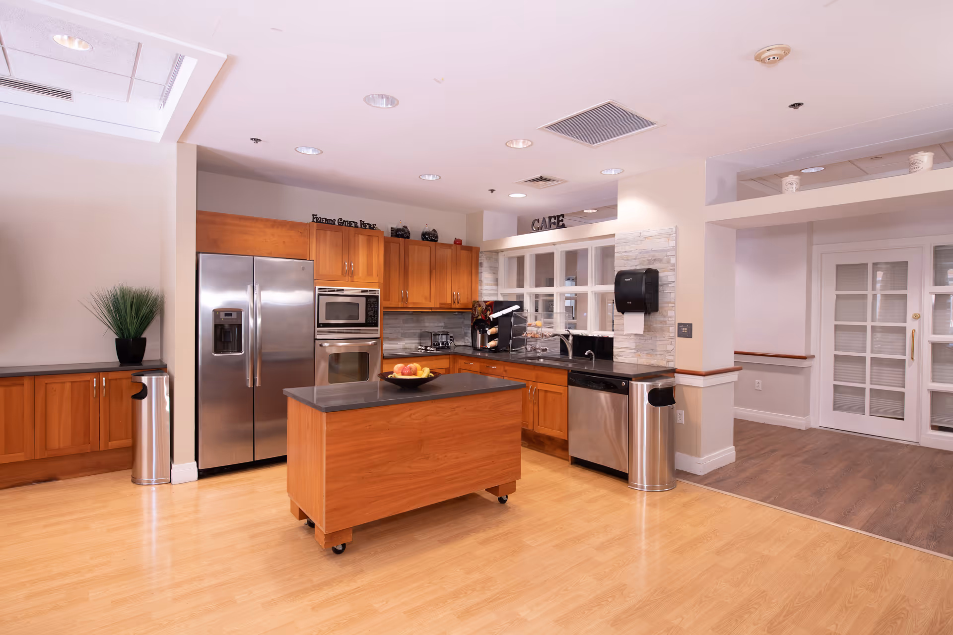 A bright communal kitchen/café with stainless steel appliances, wooden cabinets, a movable island with a fruit bowl, and countertops for serving.