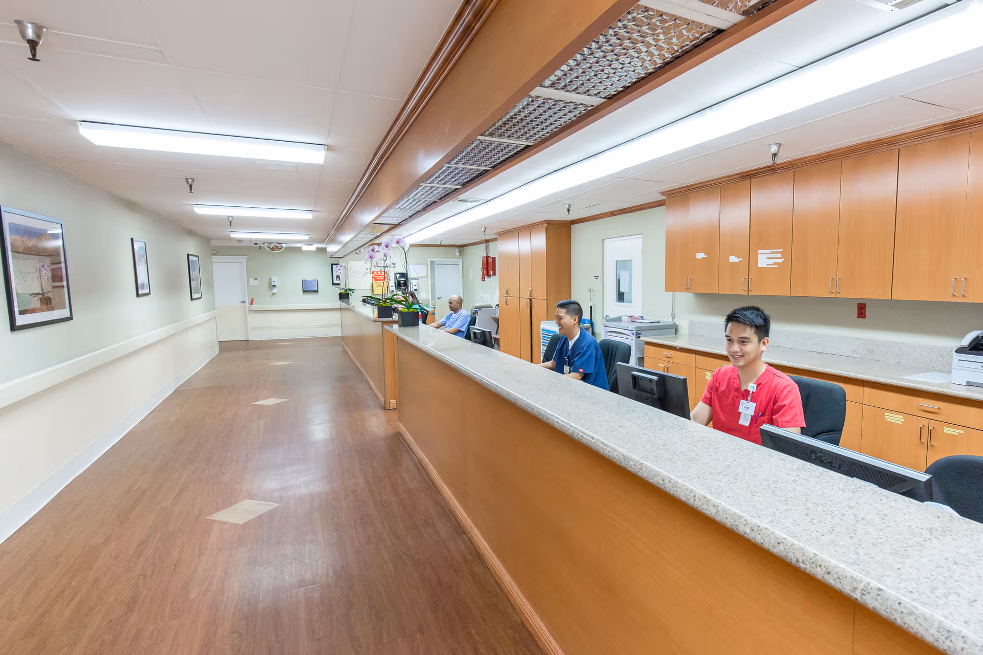 A long reception desk area in a senior care facility with three staff members seated behind the counter. The space has wooden flooring, light-colored walls with framed pictures, overhead fluorescent lighting, and wooden cabinets behind the desk.