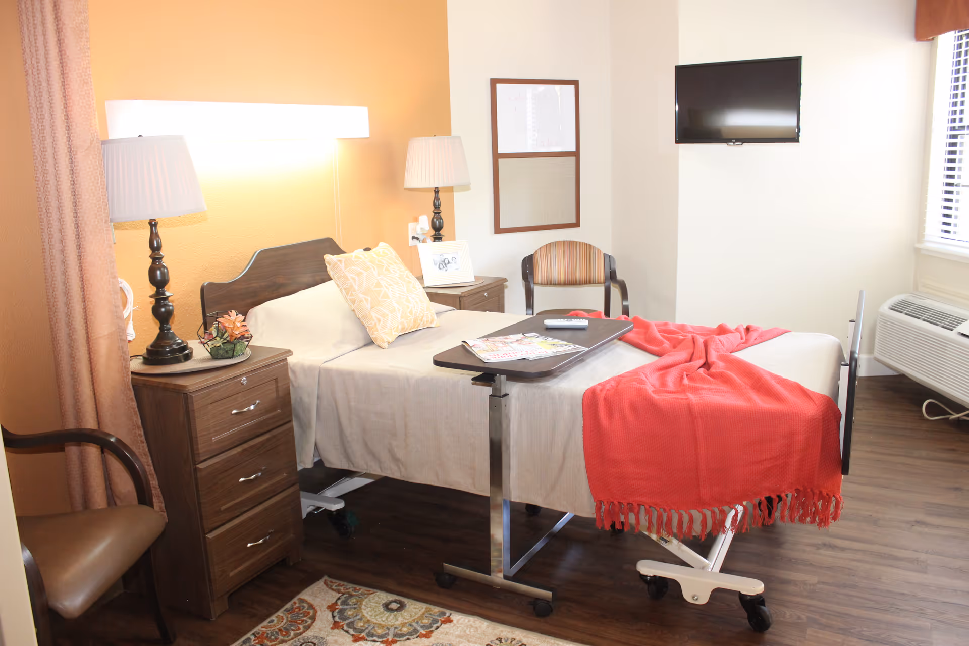 A nursing facility bedroom with a hospital-style bed covered with beige sheets and a red blanket. There is a wooden nightstand with a lamp and a small plant, a chair with a brown cushion, a wall-mounted TV, and a window with blinds. The floor is wooden, and there is a small table on wheels positioned over the bed with magazines and a remote control on it.