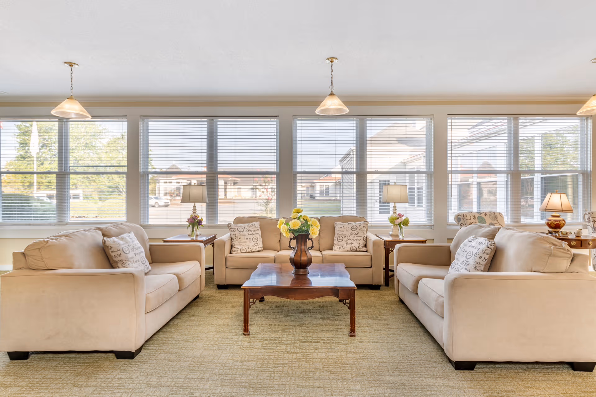 A bright and spacious living room with three beige sofas arranged around a wooden coffee table with a vase of yellow flowers. The room has large windows with blinds letting in natural light, two side tables with lamps and flower arrangements, and pendant lights hanging from the ceiling.