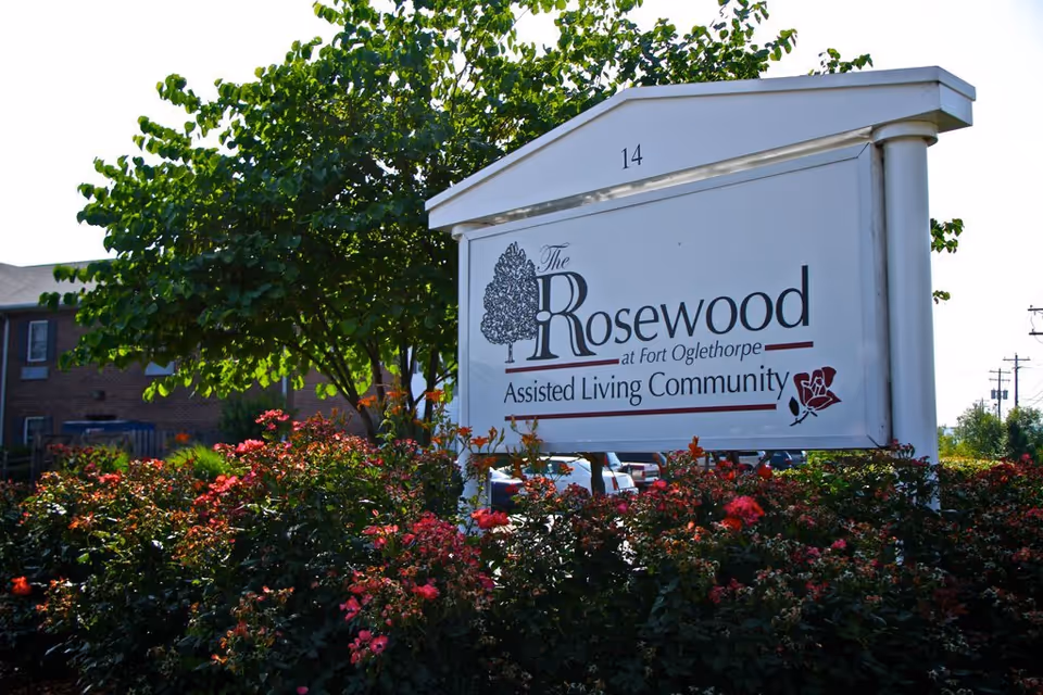 Outdoor entrance sign reading "The Rosewood at Fort Oglethorpe Assisted Living Community" surrounded by flowering bushes and a tree with part of the building visible behind.
