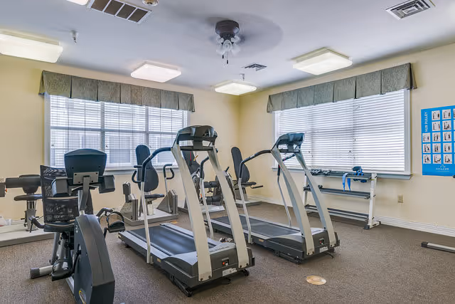 A small fitness room with two treadmills, a stationary bike, and other exercise equipment. The room has beige walls, two large windows with blinds and green valances, and ceiling lights.