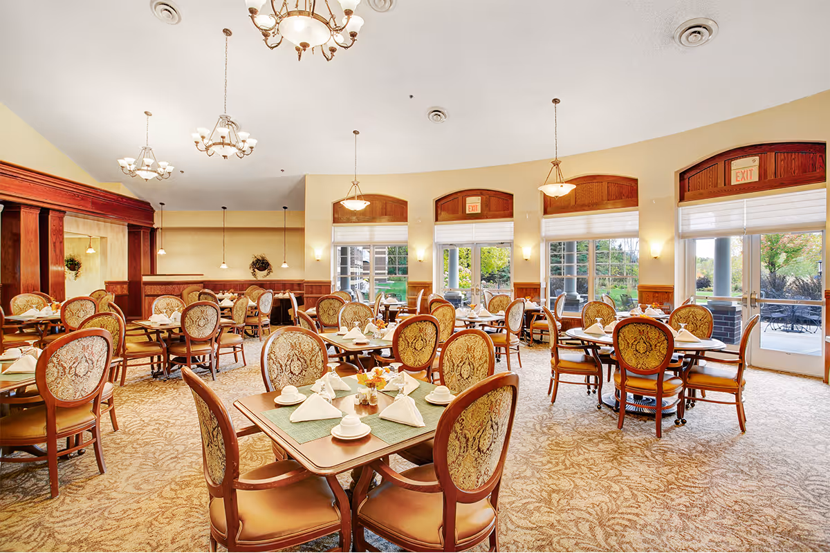 A spacious dining room with multiple wooden tables and upholstered chairs arranged neatly. Each table is set with white napkins, cups, and small flower arrangements. Large windows and glass doors allow natural light to fill the room, and elegant chandeliers hang from the ceiling. The room features warm tones with wood paneling and patterned carpet.