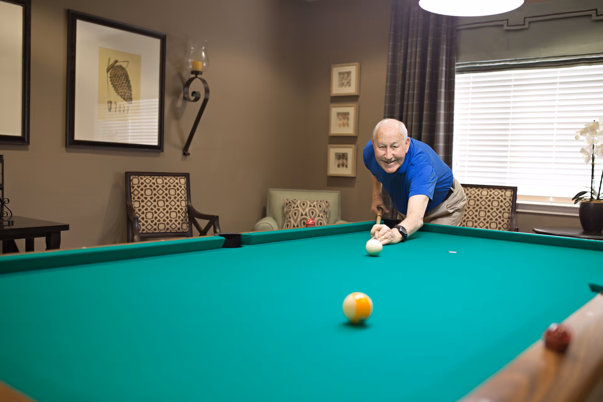 An elderly man in a blue shirt playing pool in a cozy room with patterned chairs, framed artwork on the walls, and a window with blinds and curtains.