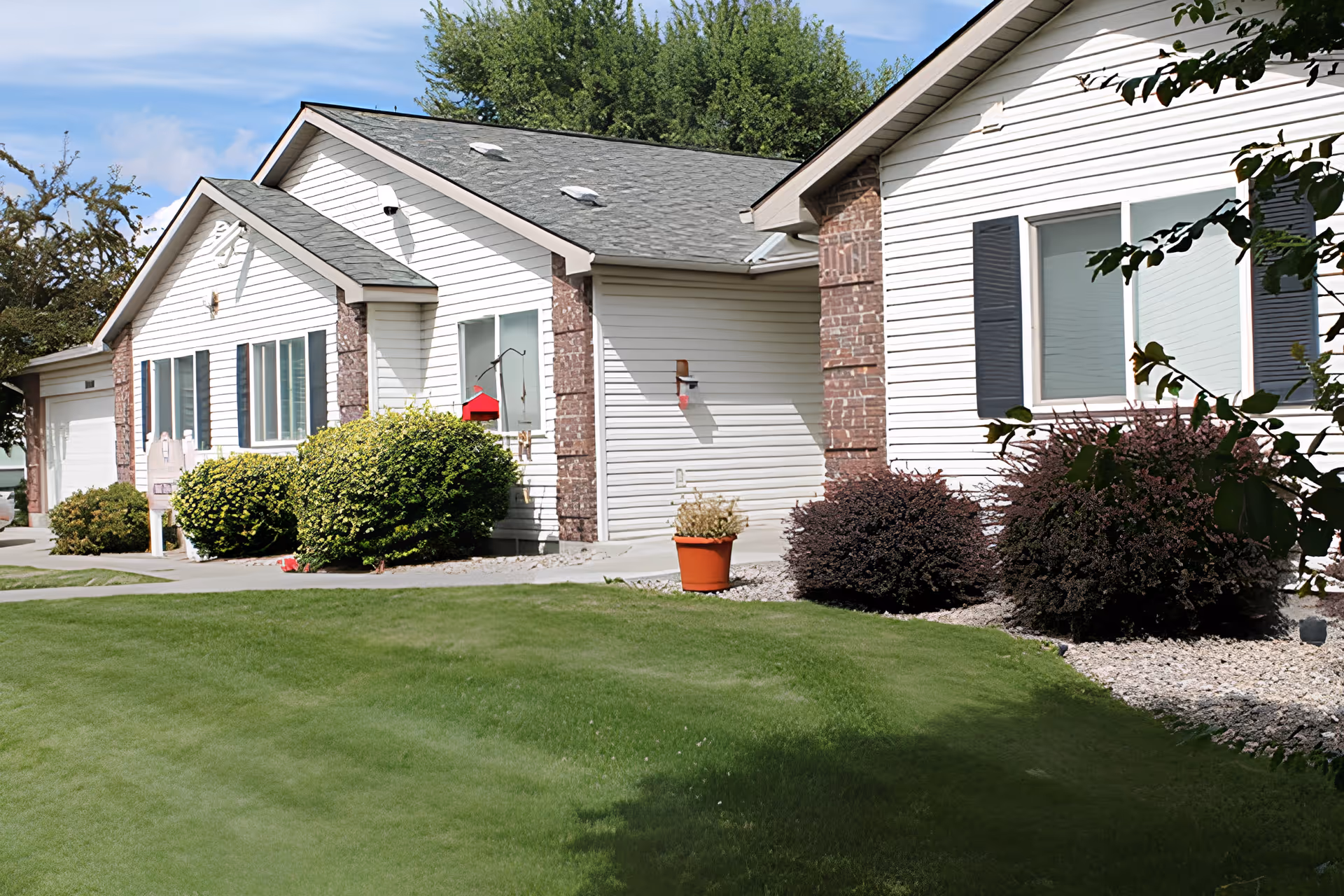 Exterior view of a single-story senior living facility building with white siding and brick accents, surrounded by green grass, bushes, and a potted plant under a partly cloudy sky.