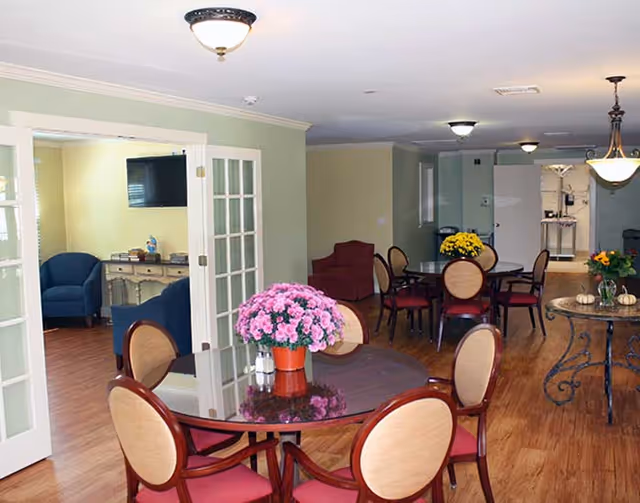 Interior view of a senior living facility dining area with round tables and chairs. Each table has a floral centerpiece with pink, yellow, or orange flowers. The room has wooden flooring, soft green and beige walls, and ceiling light fixtures. A separate seating area with blue armchairs and a TV is visible through glass-paneled double doors.