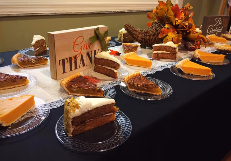 A table set with multiple slices of pumpkin pie, pecan pie, and carrot cake on clear glass plates. In the center, there is a wooden sign that says 'Give Thanks' and a decorative cornucopia filled with autumn leaves and small pumpkins. Another sign in the background reads 'Be Grateful'.