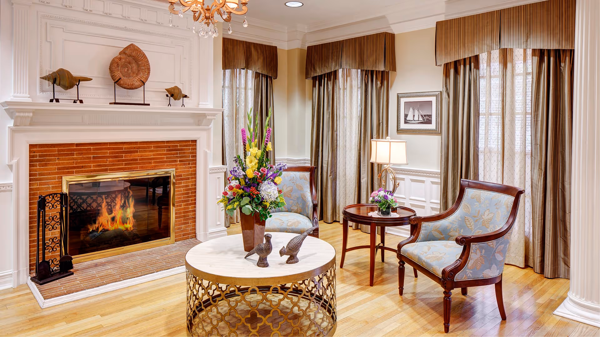 A cozy living room with a lit fireplace surrounded by a white mantel and brick interior. In front of the fireplace is a round coffee table with a decorative metal base, holding a vase with colorful flowers and two bird figurines. Two upholstered armchairs with wooden frames are positioned around the table. A small round side table with a lamp and a small flower arrangement is between the chairs. The room has hardwood floors, tall windows with sheer and brown curtains, and a framed picture on the wall.